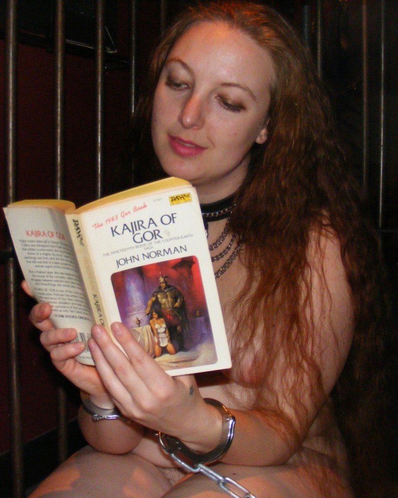 Nude, chained and collared slave girl in cage reading Kajira of Gor.